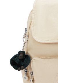 Zaino in tessuto beige con patta, dotato di due tasche con zip. Include un portachiavi a forma di gorilla in peluche verde scuro, attaccato da una catena metallica.