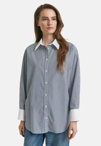 Chemise boutonnée rayée en navy et blanc, avec un col classique, des manches longues avec des poignets blancs et une coupe décontractée. Texture de tissu douce.