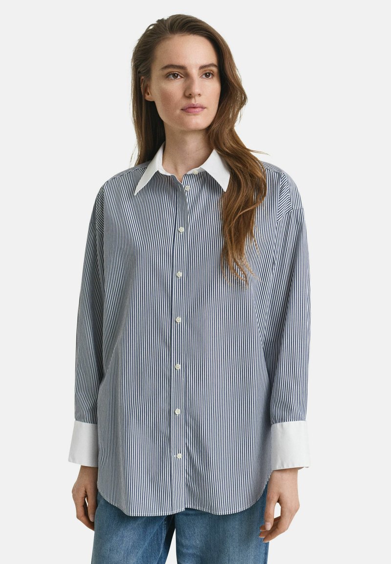 Chemise boutonnée rayée en navy et blanc, avec un col classique, des manches longues avec des poignets blancs et une coupe décontractée. Texture de tissu douce.