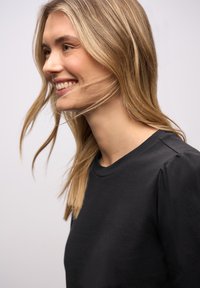 Schwarzes T-Shirt mit Rundhalsausschnitt und aufgepufften Ärmeln, mit einer glatten Textur. Das Haar des Models ist locker gestylt und erzeugt Bewegung.