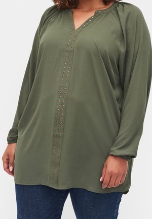 Curvy woman wearing olive green long-sleeve blouse with bronze studs along V-neckline and center front, paired with dark blue jeans.
