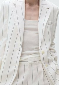 Costume en tissu rayé blanc avec un blazer structuré et un pantalon assorti. Tissu doux et texturé, présentant des rayures gris clair et des boutons discrets.