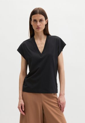Vrouw met steil bruin haar, gekleed in een zwart T-shirt met V-hals en korte mouwen en losse bruine broek, staand tegen een effen achtergrond.