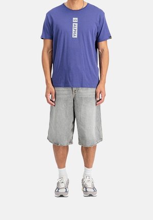 Homme portant un t-shirt bleu avec texte vertical "ALPHA", short en denim gris clair jusqu'aux genoux, chaussettes blanches et baskets grises debout.