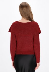 Pull en tricot rouge avec un col large à côtes et un tissu texturé. Présente un détail scintillant et des poignets ajustés, se terminant à la taille.