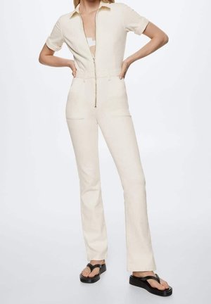 Kvinde iført cremefarvet kortærmet jumpsuit med lynlås foran, stående med hænderne på hoften og sorte flade platformsandaler.