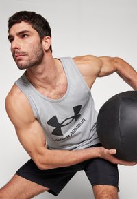 Szürke sportos ujjatlan trikó fekete "Under Armour" logóval, fekete rövidnadrággal párosítva. A férfi egy fekete, texturált medicinlabdát tart, bemutatva izmos testalkatát.