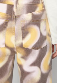 Jeans de denim desbotados com um padrão em espiral nas tonalidades de amarelo, castanho e rosa claro; apresenta costura em contraste e dois bolsos traseiros.