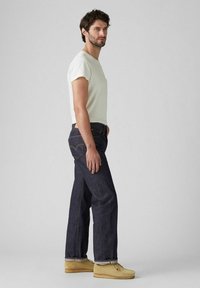 Camiseta blanca de manga corta, jeans azul oscuro con costuras en los bolsillos traseros y zapatos beige. Está de lado contra un fondo neutro.