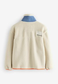 Veste en polaire crème avec un col bleu. Présente un ourlet orange et un logo brodé dans le dos. Texture confortable, coupe décontractée.