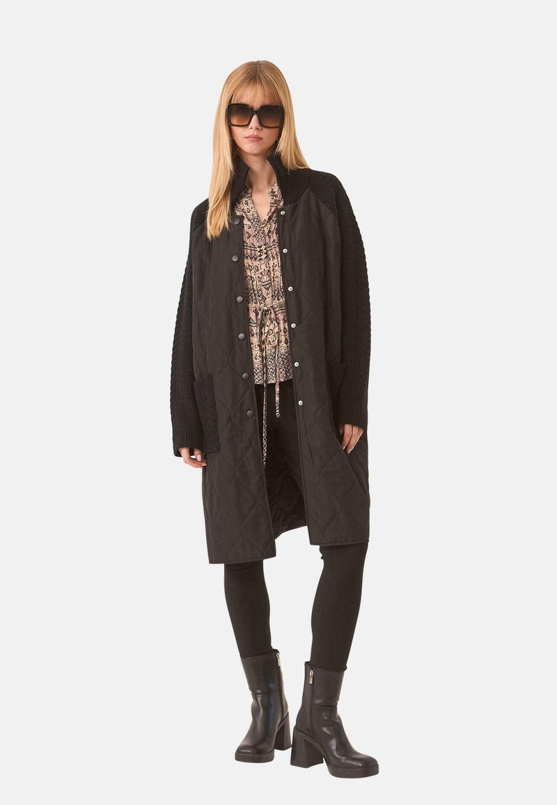 Abrigo largo acolchado negro con mangas de punto texturizado, botones de presión y dos bolsillos frontales, llevado sobre una blusa estampada y leggings negros.
