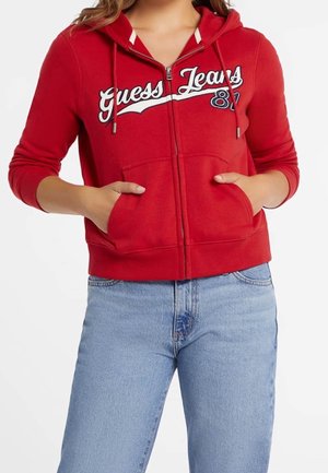 Kobieta ubrana w czerwoną bluzę z kapturem na zamek błyskawiczny z napisem „Guess Jeans 81” oraz jasnoniebieskie dżinsy, ręce w przednich kieszeniach.