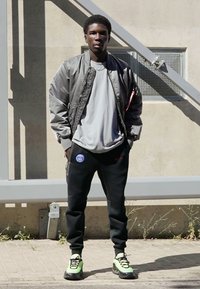 Grå bomberjakke, lys grå t-shirt, sorte joggingbukser med logo og limegrønne sneakers. Står mod en neutral baggrund.