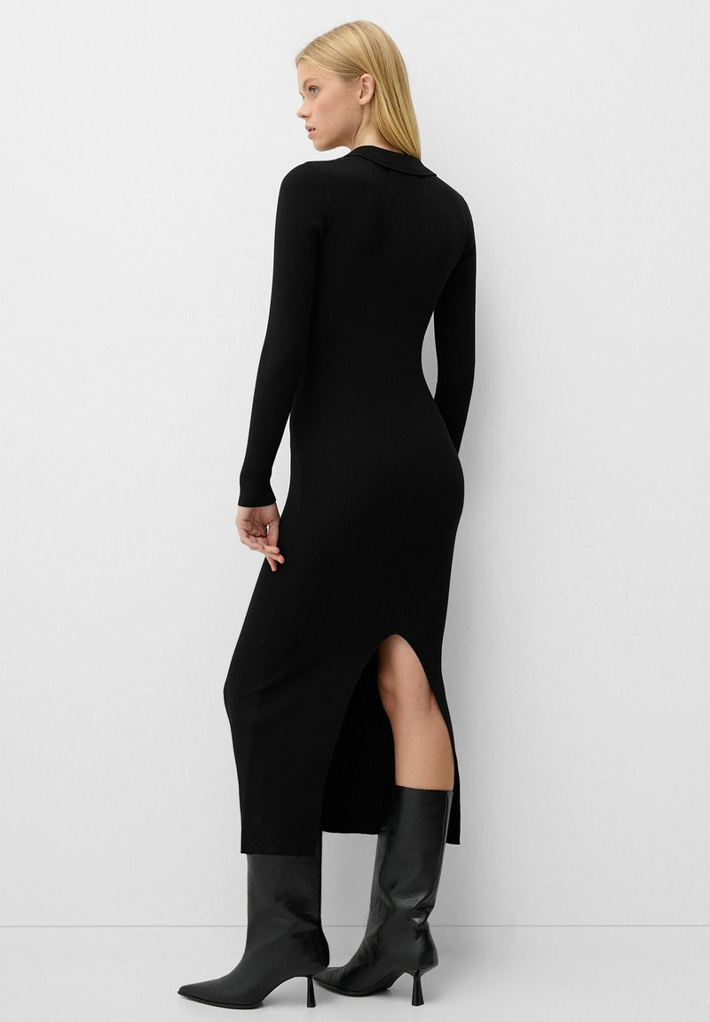 Abito in maglia a coste nero con maniche lunghe, uno spacco laterale e dettaglio colletto, abbinato a stivali neri alti fino al ginocchio con punta a forma di punta.
