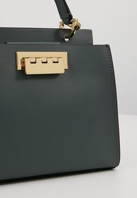Mørkegrøn lædertaske med lukning i rektangulær guldfarvet metal og kort hank, placeret mod en lys grå baggrund.