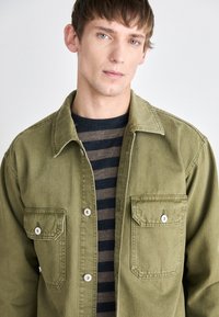 Oliven grønn denimskjorte med knapper på kragen, to brystlommer og sølvknapper, lagd over en mørk stripete t-skjorte.