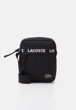 Sac à bandoulière en tissu noir avec le logo LACOSTE, fermeture éclair, sangle ajustable et logo crocodile vert brodé à l'avant.
