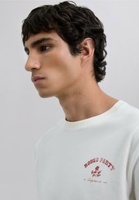 Camiseta de algodón blanca con un diseño impreso en rojo que incluye el texto "RODEO PARTY" y acentos florales, cuello redondo y mangas cortas.