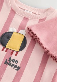 Lyserød stribet sweatshirt med en sort og gul bi-applikation med hvide vinger samt en rynket lyserød ærmedetalje. Teksten lyder "vær glad som en bi."