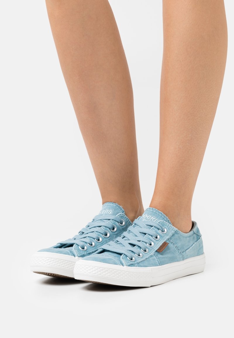 Pieds portant des baskets basses décontractées bleu clair avec des semelles et des lacets blancs, posés sur une surface grise claire unie.