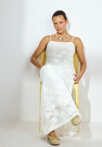 Witte gebreide jurk met spaghettibandjes, voorzien van een textuurpatroon. Draagt een chunky parelketting en lichtgekleurde schoenen. Zittend op een gele stoel.
