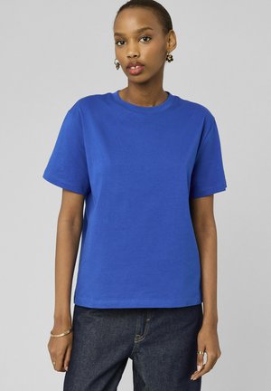 Jeune femme portant un t-shirt uni bleu roi à manches courtes et un jean en denim foncé, debout devant un fond gris clair.