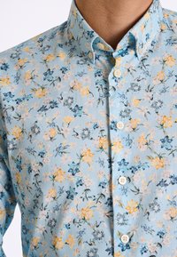 Ljusblå skjorta med knappar som har ett blommönster med gula, vita och blå blommor, tillverkad av ett mjukt tyg med en texturerad yta.
