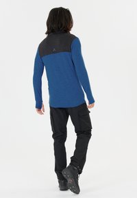 Blå långärmad topp med en svart panel på axlarna, kombinerad med svarta cargobyxor och robusta svarta skor. Ytan verkar slät.