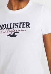 Bílý bavlněný tričko s vyšitým nápisem "Hollister California" v námořnické modré a vínové barvě, s malým černým ptáčkem pod textem.
