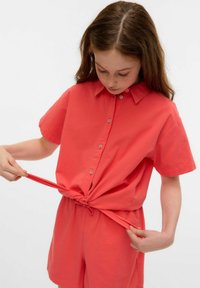 Camicetta corallo a maniche corte con bottoni e vita con coulisse. Tessuto leggero, vestibilità ampia, con una caratteristica di arricciatura regolabile nella parte inferiore.