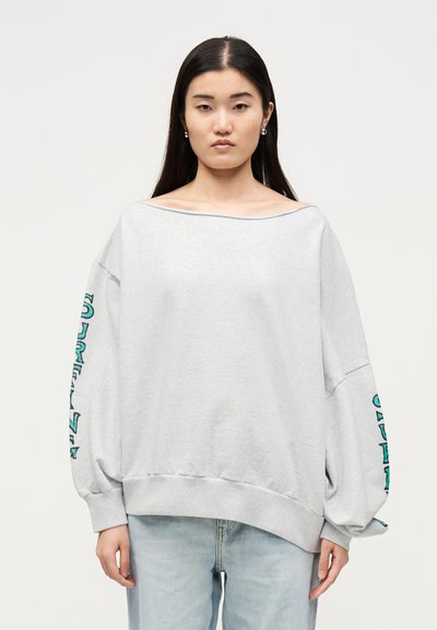 Νέα γυναίκα με μακριά σκούρα μαλλιά φοράει ένα φαρδύ ανοιχτό γκρι φούτερ με τιρκουάζ γράμματα στα μανίκια και ανοιχτό μπλε τζιν, στέκεται μπροστά σε λευκό φόντο.