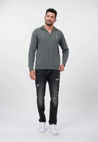 Grau gestrickter Pullover mit Reißverschluss am Kragen, versehen mit einem Zopfmuster. Kombiniert mit zerrissenen schwarzen Jeans und weißen Sneakern.