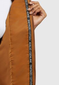 Giacca isolante marrone con zip frontale. L'interno presenta una rifinitura in tessuto nero con marchio bianco. Texture liscia e vestibilità aderente.