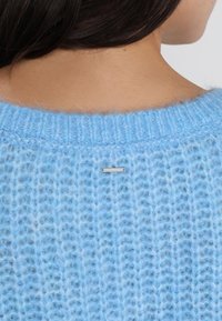 Ljust blått stickat tröja med en lös, texturerad design. Har en liten metallisk etikett nära halsringningen. Mjuk och luftig framtoning.