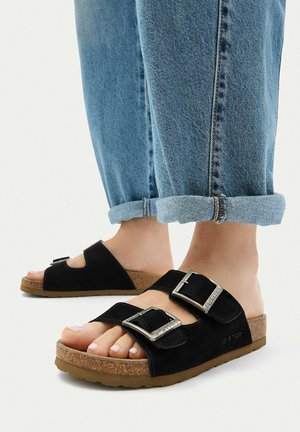 Piedi con sandali in suede nero con doppia fibbia argentata e suole in sughero, abbinati a jeans di denim blu risvoltati.