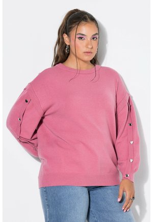 Kvinde iført en lyserød sweater med hjerteformede knapper på ærmerne og blå jeans, stående mod en hvid baggrund.