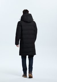 Abrigo de plumas negro con capucha, de longitud hasta la rodilla, con costuras horizontales y una textura suave. Se lleva con jeans azules y calzado marrón.