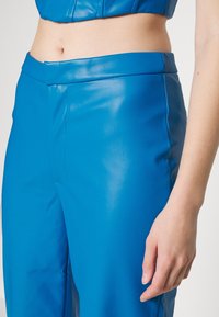 Detailní záběr na osobu v jasně modrých, vysokých umělých kožených kalhotách s hladkým povrchem a úzkým pasem.