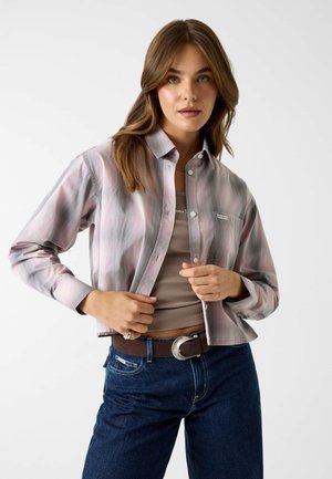 Femme portant une chemise rose clair et grise boutonnée sur un crop top beige, un jean bleu et une ceinture marron avec une boucle argentée.