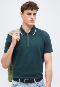 Νέος άνδρας που φοράει σκουρό πράσινο κοντομάνικο πόλο με φερμουάρ στο γιακά, ανοιχτό μπλε τζιν, μαύρη ζώνη, κρατώντας πράσινο πουκάμισο με καρό πάνω στον ώμο.