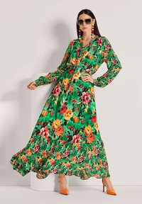 Langes grünes Blumenkleid mit lebhaften orangefarbenen, pinken und gelben Blumen. Es hat lange Ärmel, eine knöpfbare Vorderseite und einen gestuften, gerafften Saum.