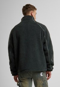 Dark green fleece jacket with high collar, contrasting white stitching along sleeves, and textured fabric. No visible branding on the back.