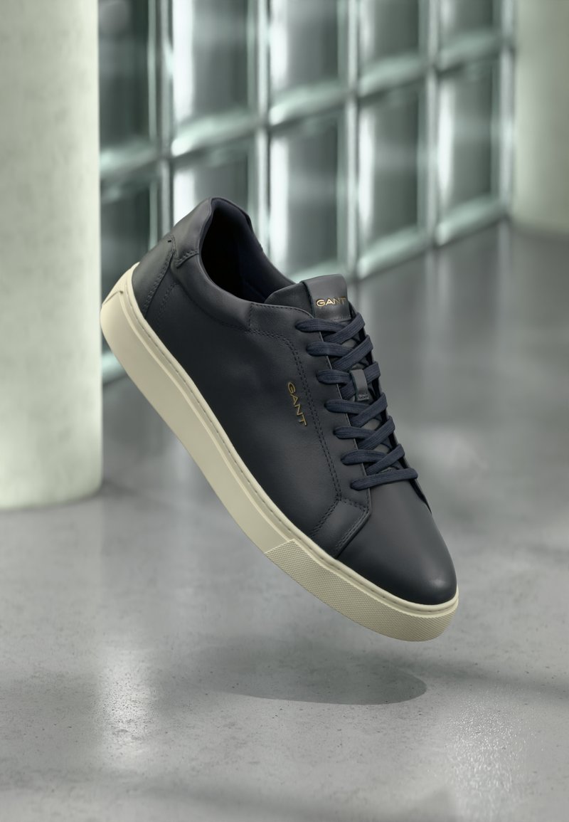 Navy leather sneaker with rounded toe, flat laces, and textured sole; features logo on tongue, contrasted by a cream-colored outsole.