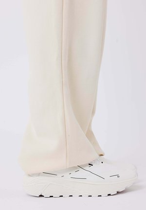 Beige weit geschnittene Hose aus weichem Stoff, kombiniert mit weißen Sneakern mit schwarzen Linienakzenten und strukturierten Sohlen.