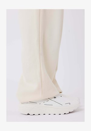 Beige weit geschnittene Hose aus weichem Stoff, kombiniert mit weißen Sneakern mit schwarzen Linienakzenten und strukturierten Sohlen.