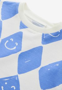 Camisa de manga corta blanca con patrones a cuadros azules y caras sonrientes, hecha de suave algodón, acabada con un cuello redondo ribeteado.