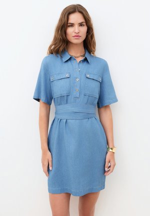 Kvinde iført en kortærmet, lyseblå denimskjortekjole med forlommer, knapper og et bredt taljebælte, stående foran en hvid baggrund.