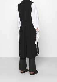 Zwarte mouwloze jurk met plooien aan de achterkant, gecombineerd met een wit shirt met lange mouwen. Het model draagt zwarte wijde broekspijpen en schoenen.