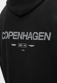 Μαύρο φούτερ με κουκούλα με λευκό ριγέ κείμενο "COPENHAGEN" και μικρότερο λευκό κείμενο που περιλαμβάνει "CPH", "ARKK COPENHAGEN" και "EST 2015" στην πλάτη.