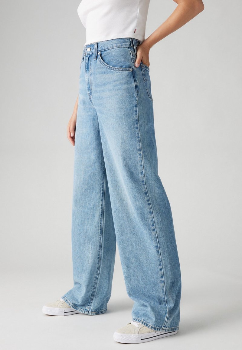 Personne portant un jean large bleu clair, des baskets blanches et un t-shirt blanc, debout avec une main dans la poche devant un fond uni.